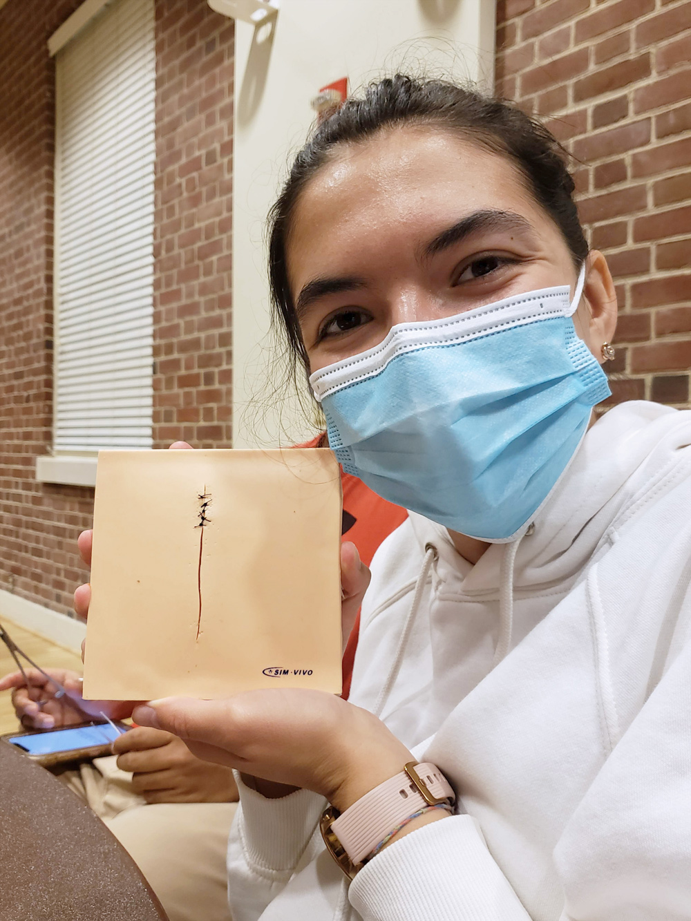 A female student wearing a surgical mask, holding up a model on which she has practiced making sutures.